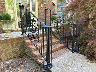 Black scrollwork porch railings beside brick steps and large planters